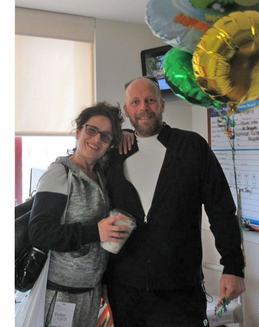 Picture shows Paul and Jomania standing near his hospital bed.  She has one arm around him and is leaning on his shoulder and smiling at us, holding a bag over her other shoulder.  He is holding the large balloons and smiling.